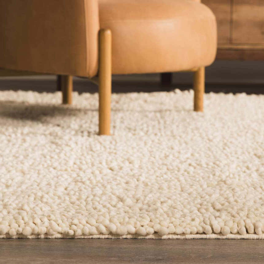 Close-up of the cream Oakridge Area Rug, a hand-woven wool rug with a textured, looped pile, under a tan leather chair. Styleshot
