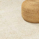 Close-up of the cream, hand-woven Oakridge Area Rug with a high-pile texture, paired with a jute pouf. Styleshot