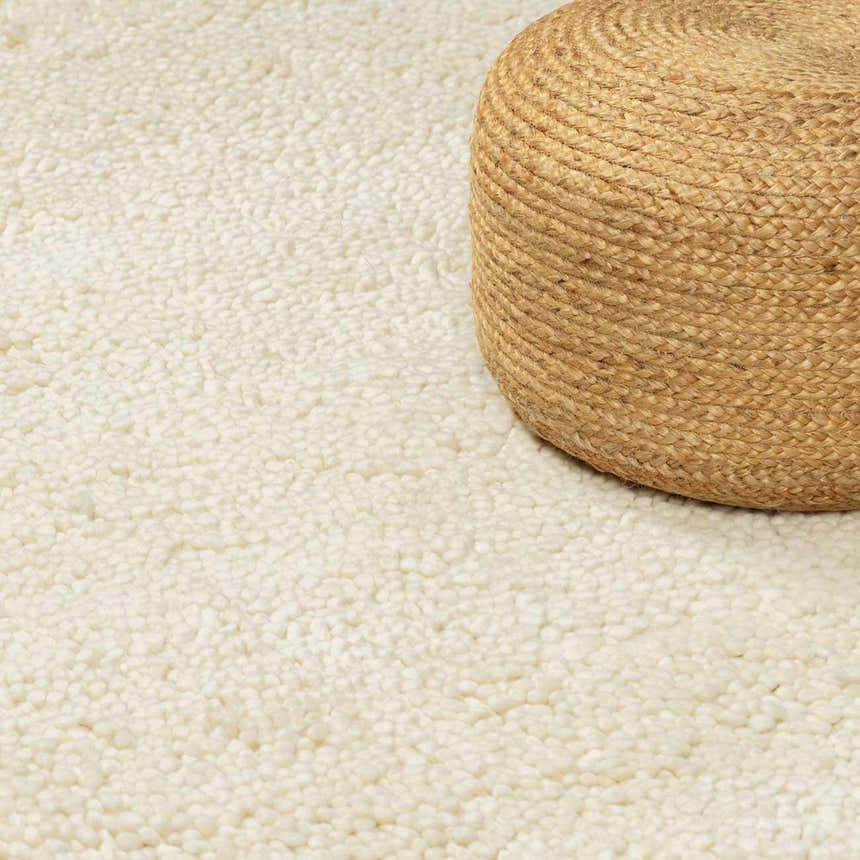 Close-up of the cream, hand-woven Oakridge Area Rug with a high-pile texture, paired with a jute pouf. Styleshot