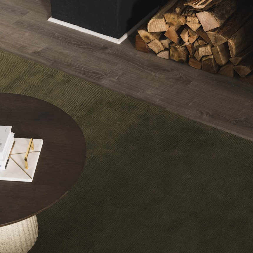 Living room scene with a solid green Brockton wool rug, round wood coffee table, and fireplace with stacked wood. Styleshot