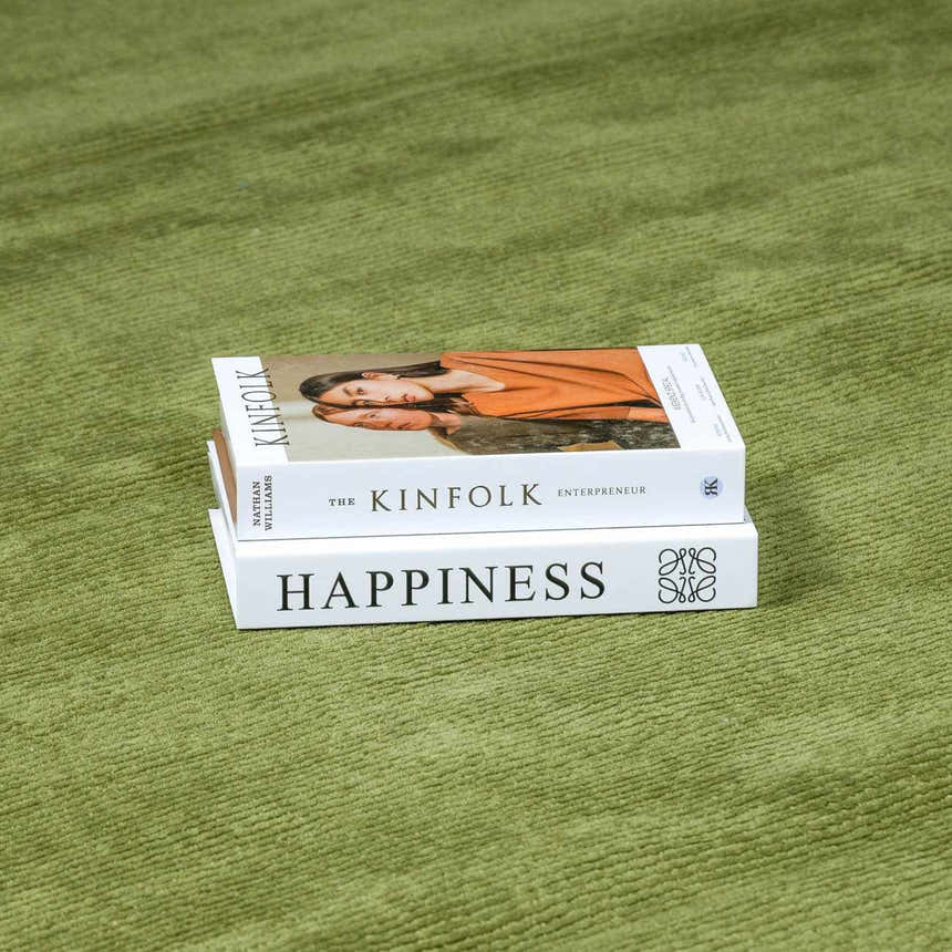 Close-up of the solid green, hand-loomed wool texture of the Brockton Area Rug, styled with stacked books. Styleshot