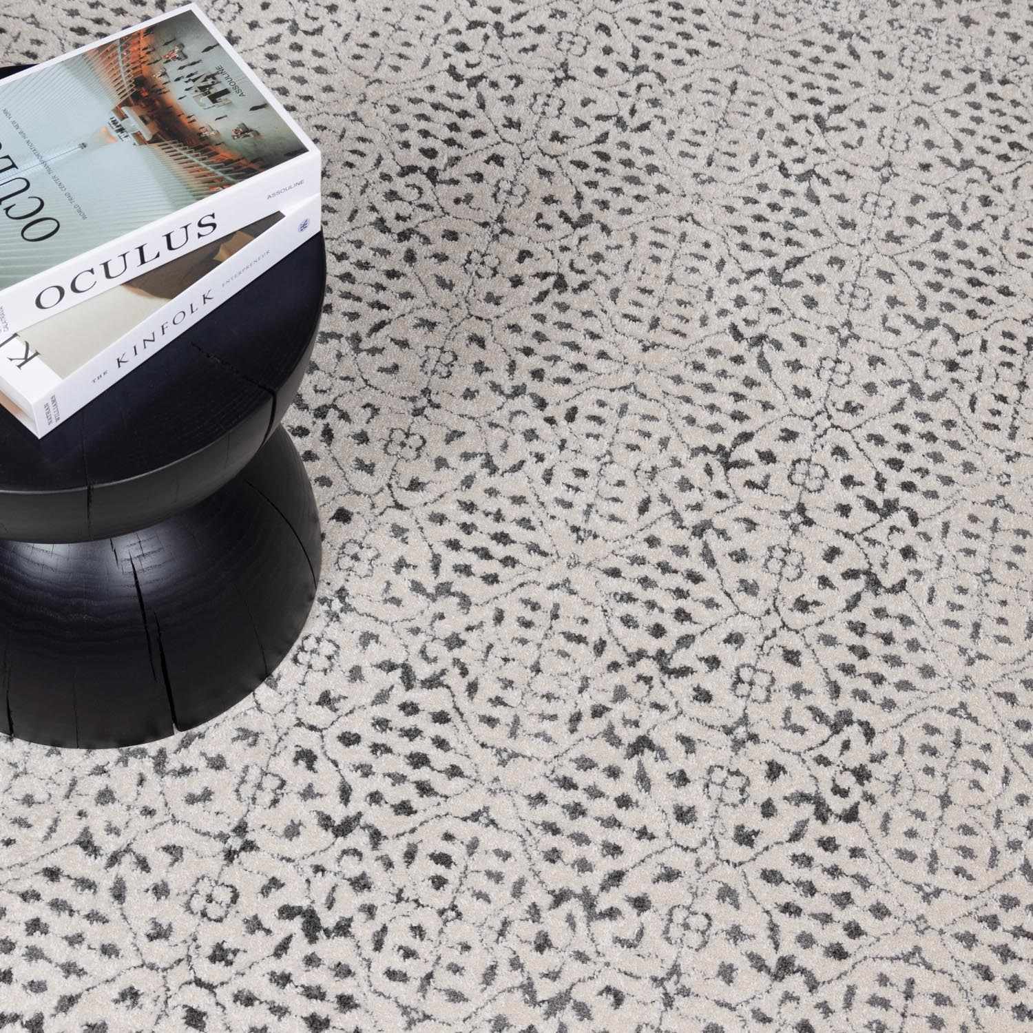 Close-up of the Sattley Area Rug. Beige rug with charcoal floral-like pattern and a black stool with books. Styleshot