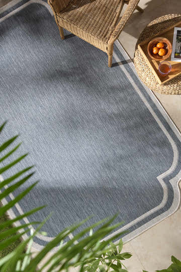 Outdoor patio scene with a blue Arnola rug, wicker chair and table, and tropical plants. Roomscene-Rectangle