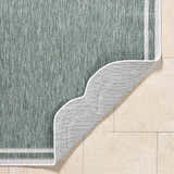 Close-up of the Arnola Green Outdoor Rug showing its texture, cream border, and non-slip backing on beige tile. Fold