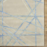 Close-up of the Hiba Area Rug showing a beige wool texture with a slate blue geometric line pattern. Corner