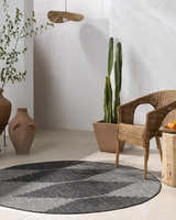 Minimalist patio scene with a round Stephan Area Rug in black and cream, wicker chair, and potted cactus. Roomscene-Round