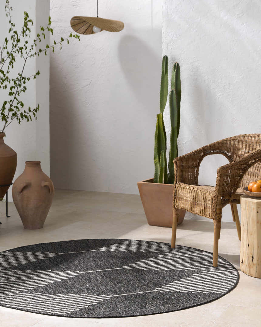 Minimalist patio scene with a round Stephan Area Rug in black and cream, wicker chair, and potted cactus. Roomscene-Round
