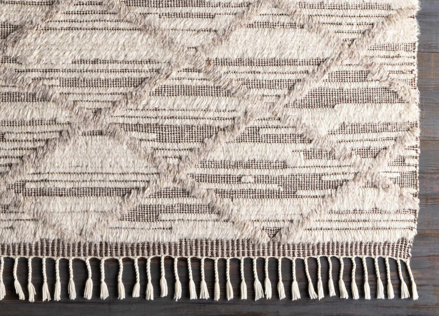Close-up of the Tankerton Area Rug. Oatmeal and dark brown wool rug with a textured, geometric pattern and fringe. Corner