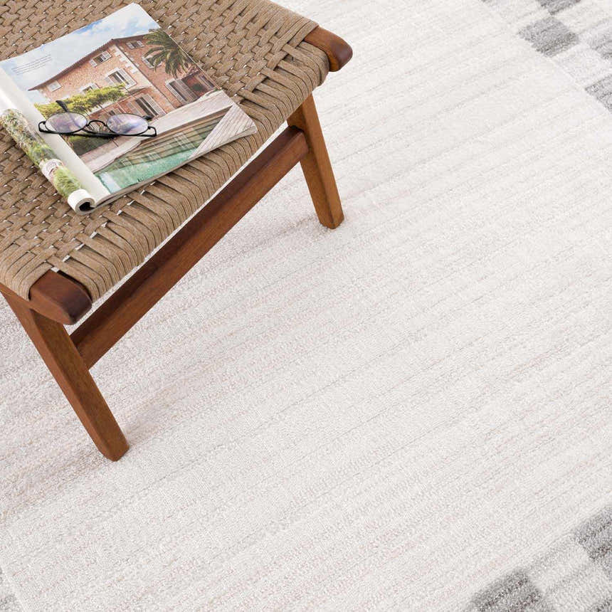 Close-up of the Azrika Gray Checkered Washable Rug with beige and gray tones, next to a wooden stool. Styleshot
