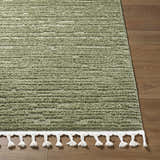 Close-up of the olive green Annily Area Rug showing its texture and white braided fringe on a wood floor. Front