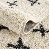 Close-up of the Alstead Shag Area Rug, showing its beige shag texture and charcoal Moroccan-inspired cross pattern. Fold