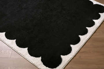 Close-up of the Pidaa Cream Border Rug. Black center with a cream, scalloped border on a wood floor. Corner