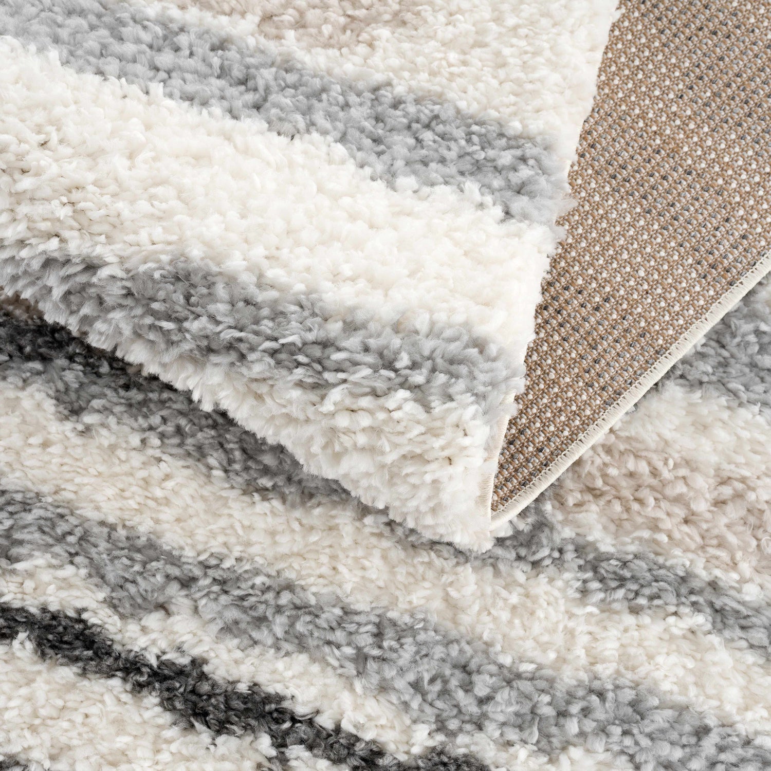 Close-up of the Zoya Irregular Striped Area Rug showing beige, cream, and gray shag stripes and woven backing. Fold