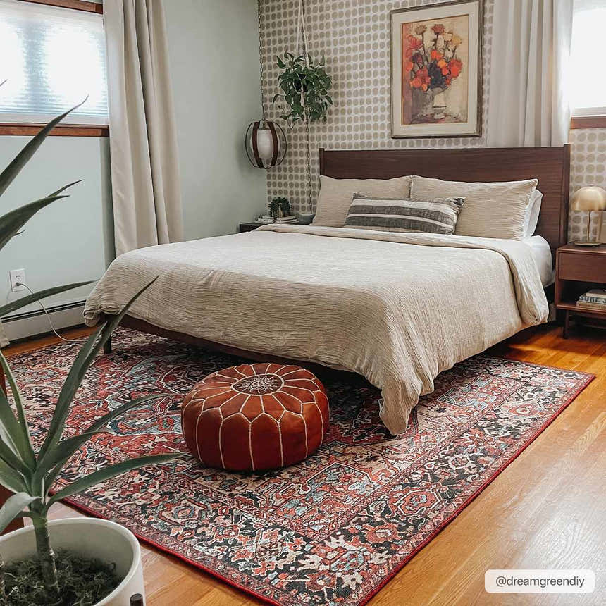 Bedroom scene with a red and navy Neyland Area Rug, beige bedding, and a brown leather pouf. Roomscene-Rectangle
