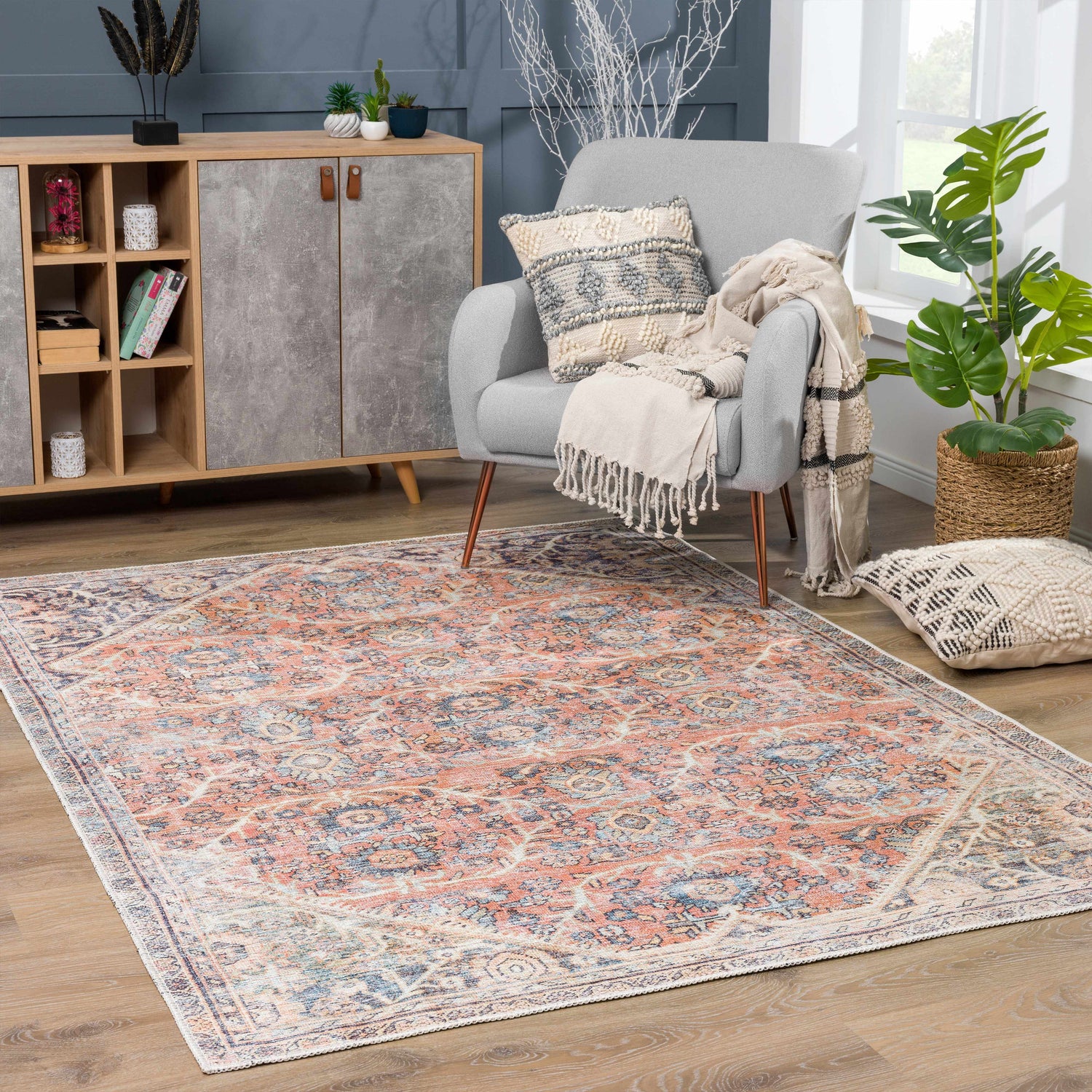 Living room scene with a Konstantina Washable Area Rug in peach, blue and beige. Gray armchair, wood cabinet, and plants. Roomscene-Rectangle