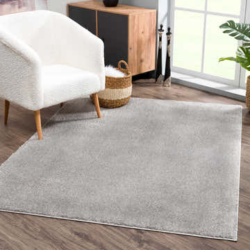 Living room scene with a Judy Solid Gray Washable Shag Rug, white armchair, wood cabinet, and a bright window. Roomscene-Rectangle