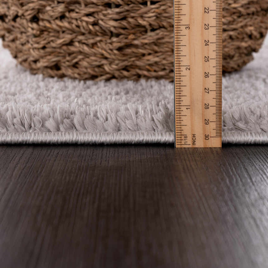 Close-up of the Judy Solid Gray Washable Shag Rug showing the pile height with a ruler and a basket on a dark wood floor. Pile