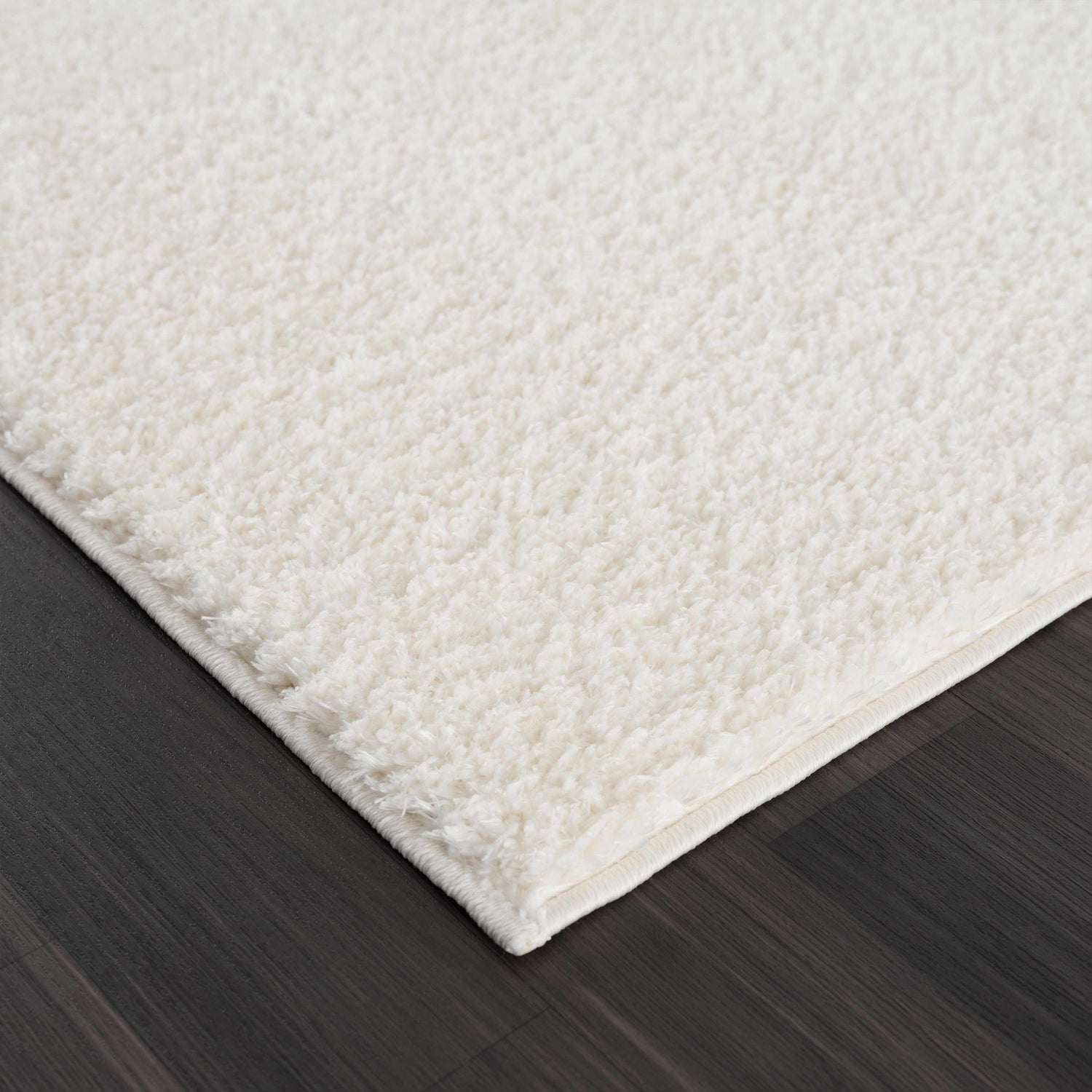 Close-up of the off-white Judy Washable Area Rug showing its plush shag texture on a dark wood floor. Corner