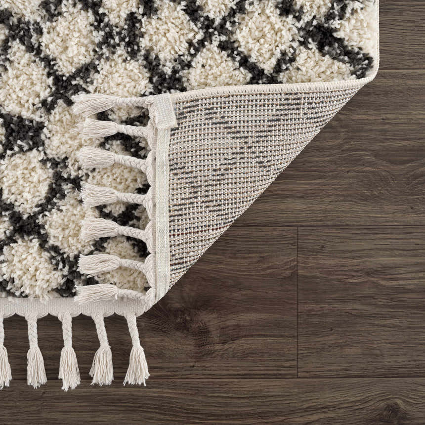 Close-up of the Muni Berber Shag Rug showing charcoal and light beige pattern, tassels, and machine-woven construction. Fold