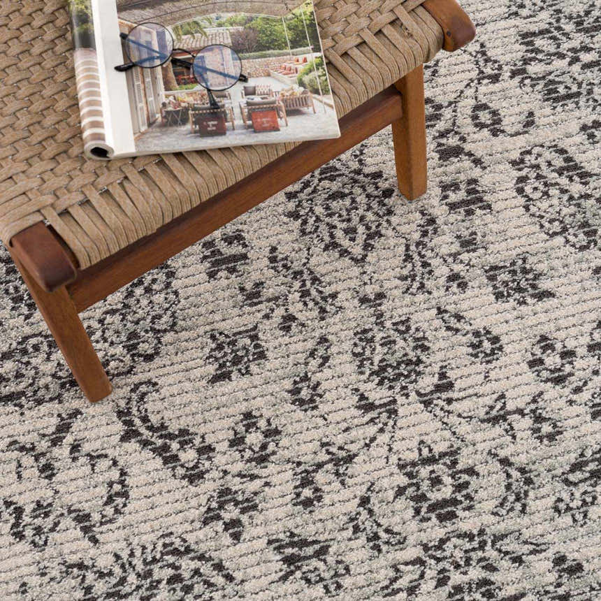 Close-up of the Paicines Area Rug showing charcoal and beige floral pattern and texture next to a woven chair. Styleshot