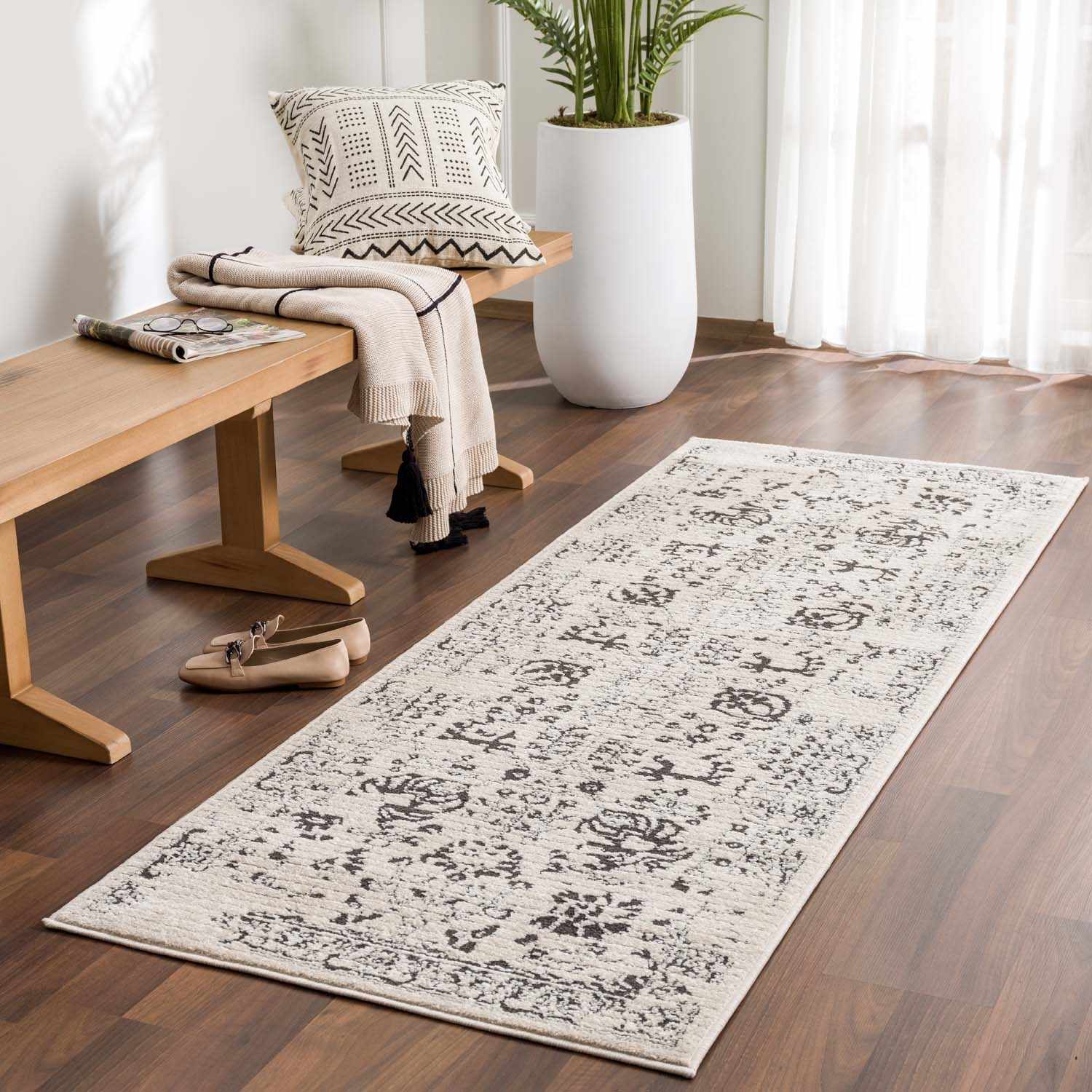 Entryway scene with a beige Tallmadge area rug, wooden bench, throw blanket, and potted plant against a white wall. Roomscene-Runner