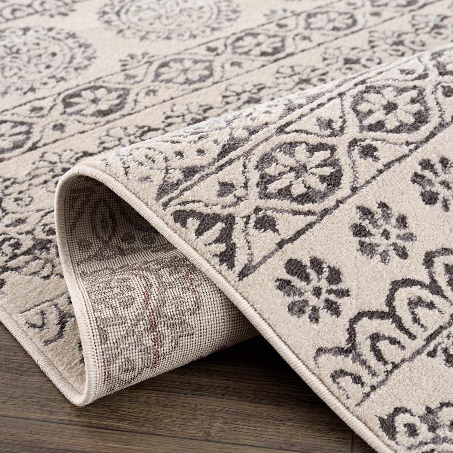 Close-up of the Warroad Area Rug. Beige rug with a charcoal, gray, and light beige traditional floral pattern. Fold