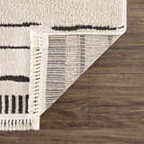 Close-up of the bone-colored Aulii Area Rug showing the texture, dark stripes, fringe, and backing on a wood floor. Fold