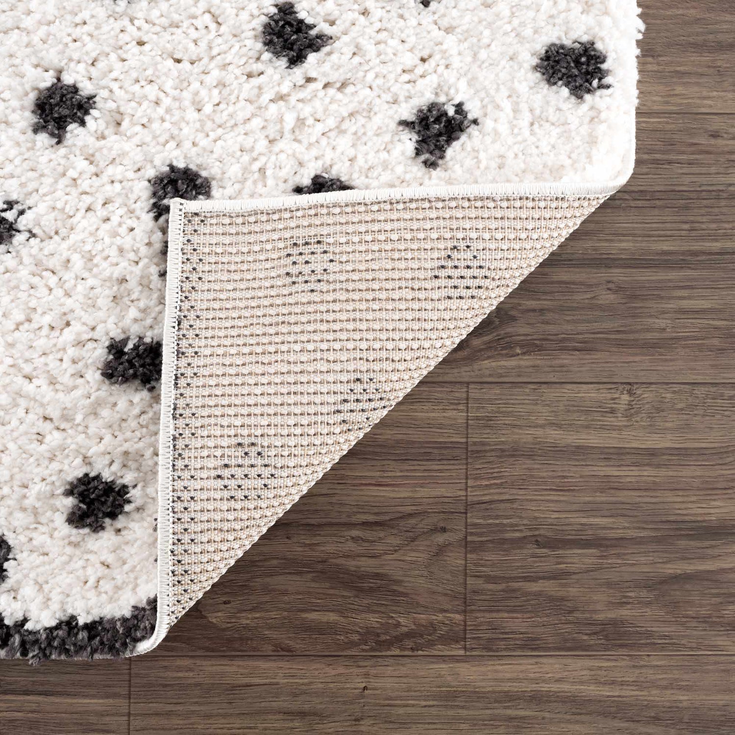 Close-up of the Chaia Dotted Black & White Plush Rug showing texture, black dots, and the rug's backing on a wood floor. Fold