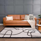 Living room scene with a tan leather sofa and the Brad Area Rug, a white shag rug with black abstract lines. Roomscene-Rectangle