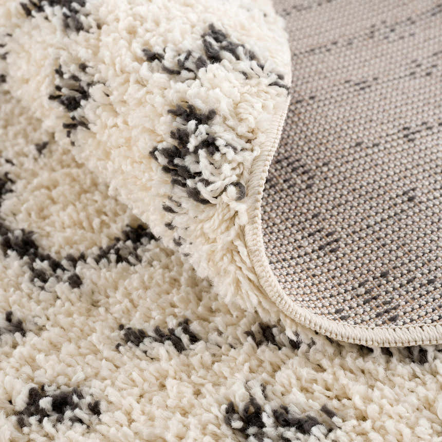 Close-up of the Kibureau Area Rug showing its beige shag texture and charcoal Moroccan-inspired pattern. Fold