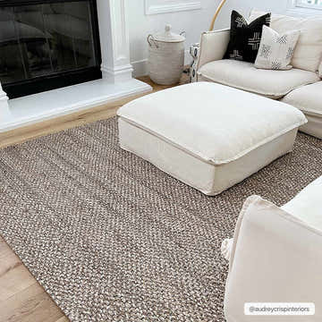 Living room scene with a Caspar Hand Woven Natural Jute Rug, cream sofa, ottoman, and fireplace. Neutral, cozy aesthetic. Roomscene-Rectangle