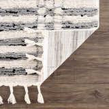 Close-up of the Nava Area Rug showing beige, black, and bone geometric pattern, tassels, and machine-woven construction. Fold