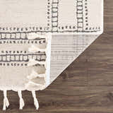 Close-up of Ryuu Ivory & Gray Area Rug showing the geometric pattern, tassels, and rug backing on a wood floor. Fold