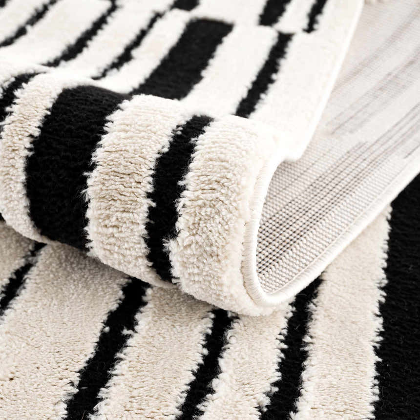 Close-up of the Bansi Area Rug showing its beige and black geometric stripe pattern and plush, machine-woven texture. Fold