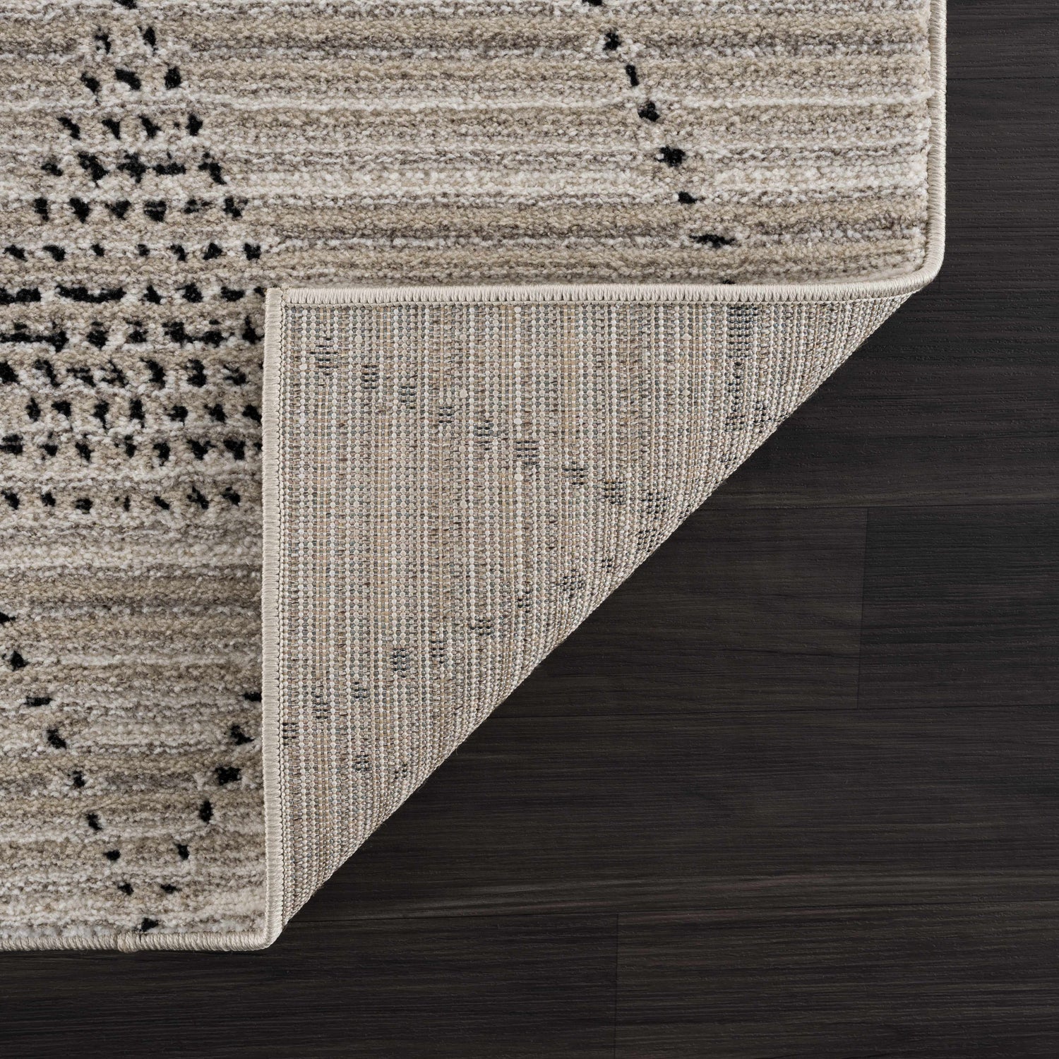 Close-up of the Lexi Area Rug. Beige and ivory rug with a geometric design on a dark wood floor. Rug backing visible. Fold
