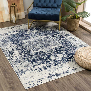 Living room scene with a Rachel Navy Area Rug, blue velvet chair, and woven pouf on wood floors. Roomscene-Rectangle