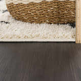 Close-up of the Alstead Shag Area Rug showing its beige color, thick shag texture, and charcoal pattern. Woven basket detail. Pile
