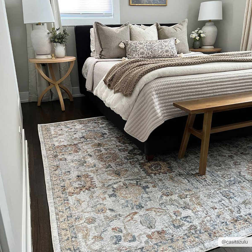 Bedroom scene with Dion Washable Floral Rug in charcoal, beige, and gray tones under a bed with a wooden bench. Roomscene-Rectangle