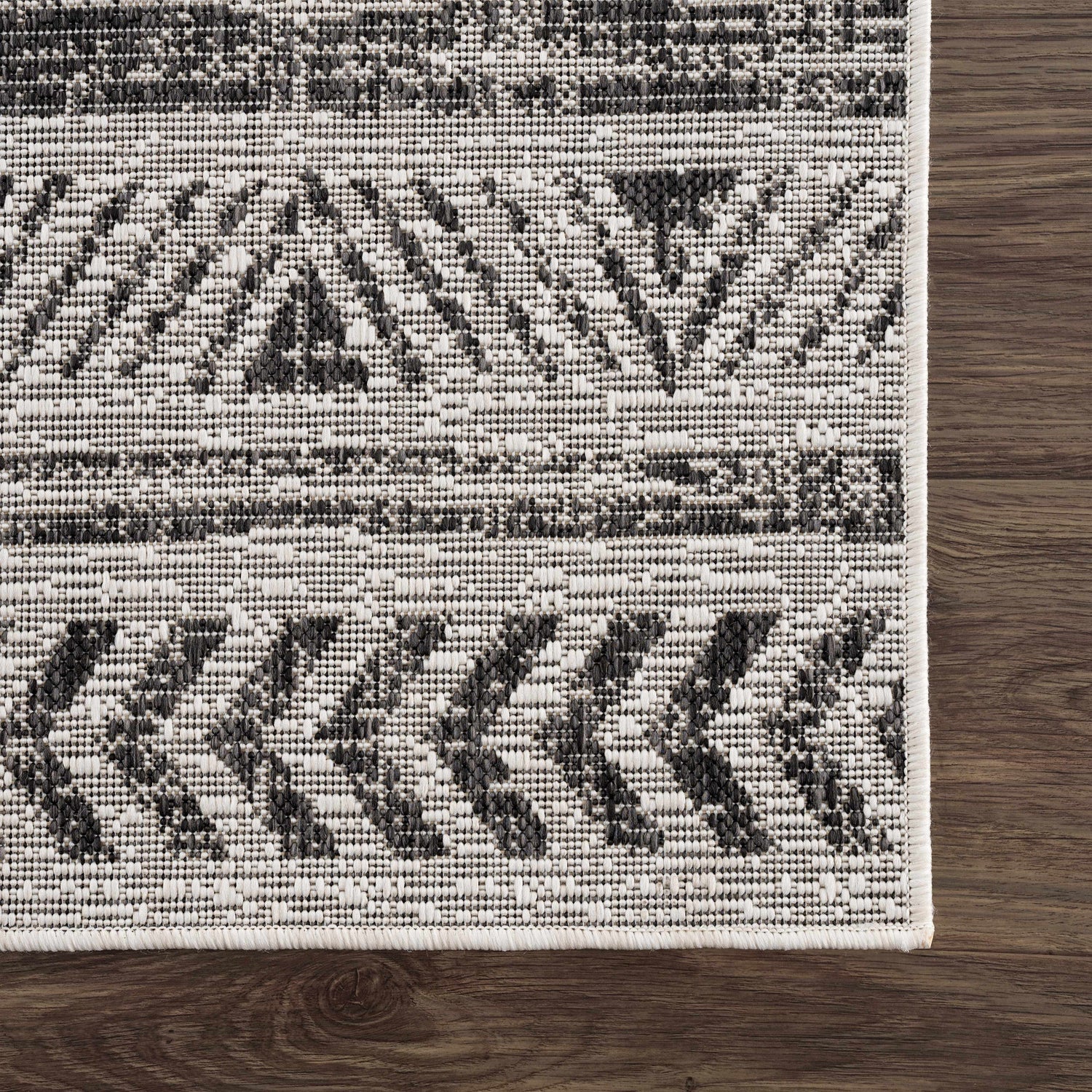 Close-up of the Catrine Indoor & Outdoor Rug. Bohemian style rug in black, charcoal, and off-white colors on dark wood floor. Swatch