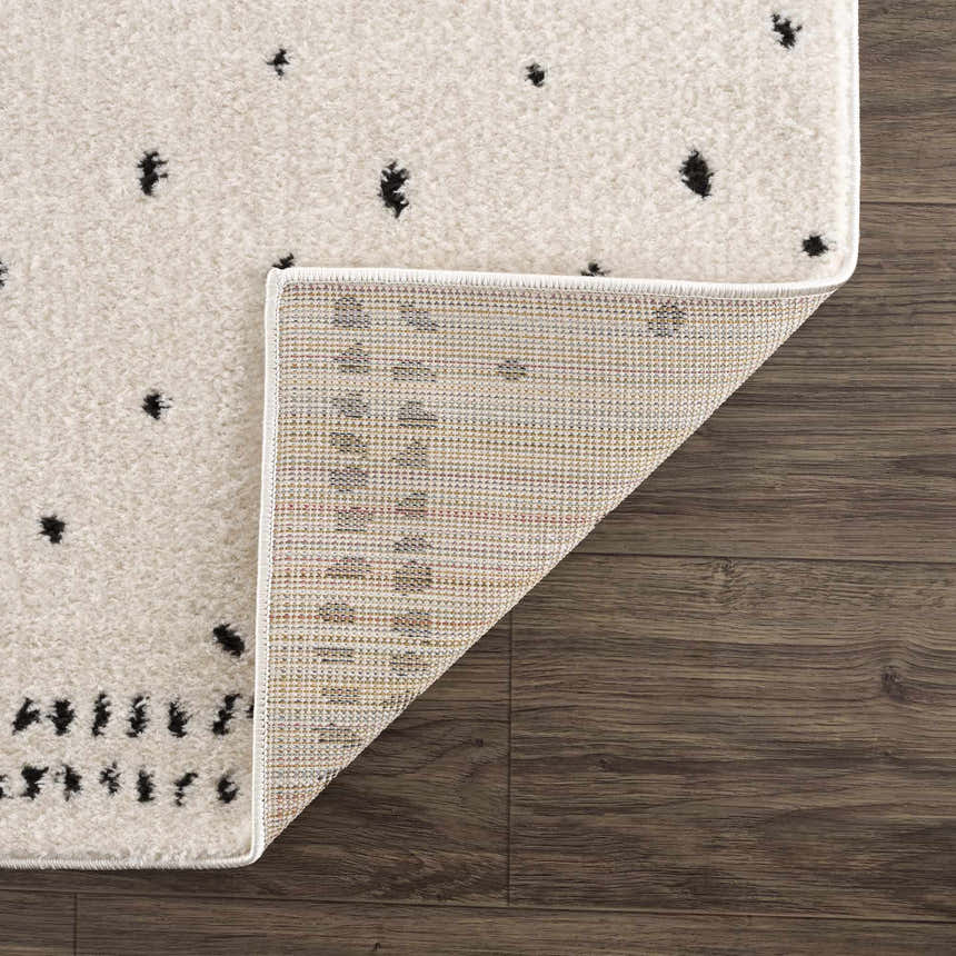 Close-up of Julie Area Rug showing beige pile with black polka dot pattern and rug backing on dark wood floor. Fold