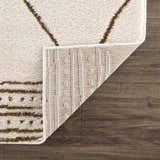 Close-up of the Keone Area Rug showing beige shag texture and brown geometric pattern with rug backing on dark wood floor. Fold
