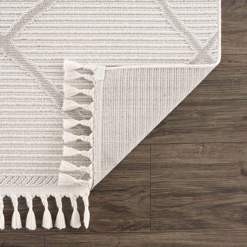 Close-up of the Lova Area Rug showing its geometric design, tassels, and the rug's backing on a dark wood floor. Fold