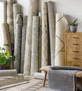 Rolled Plean Premium Rugs in neutral colors, including gray, cream, and brown, stacked in a room. Front