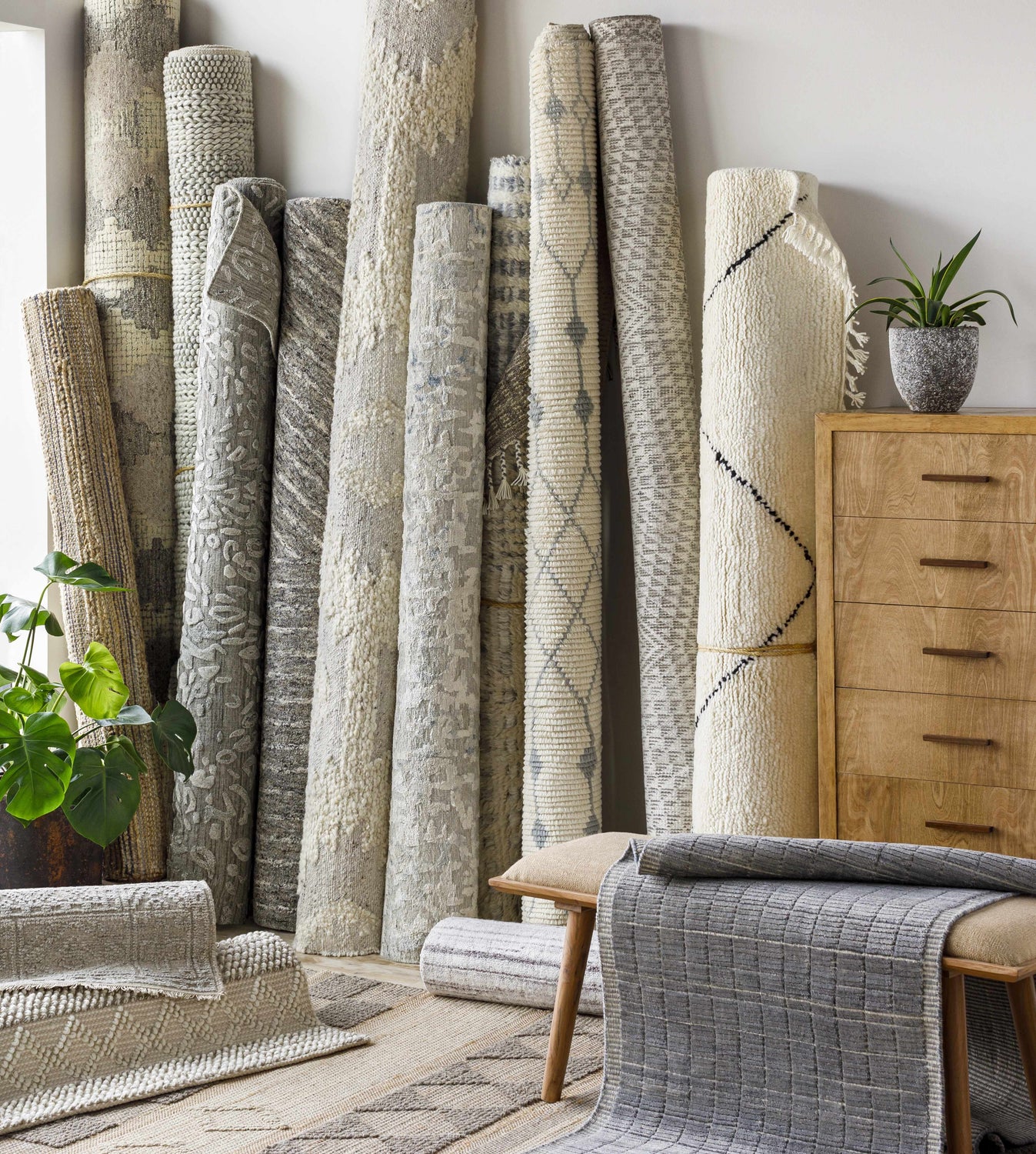 Rolled Plean Premium Rugs in neutral colors, including gray, cream, and brown, stacked in a room. Front