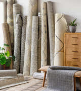 Rolled Pooleville Area Rugs in ivory and gray with various patterns, leaning against a wall with plants and a wooden dresser. Front