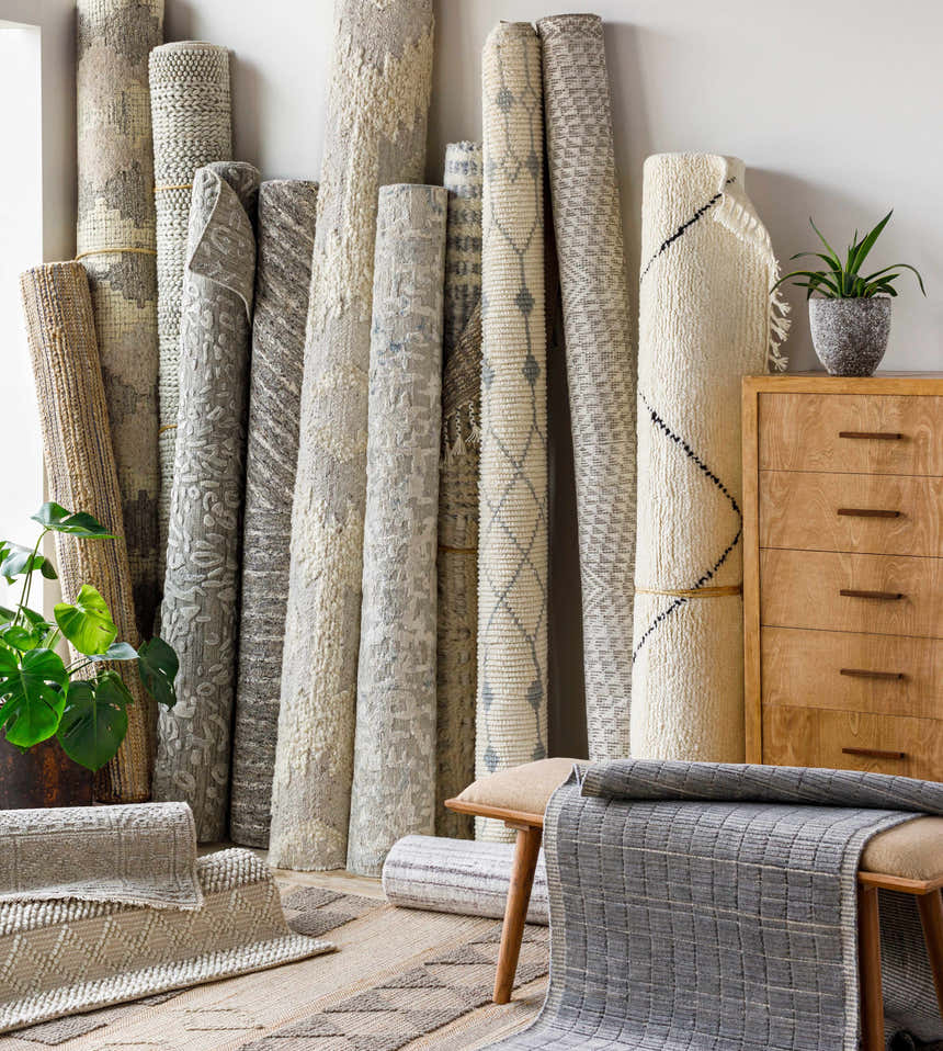 Rolled Pooleville Area Rugs in ivory and gray with various patterns, leaning against a wall with plants and a wooden dresser. Front