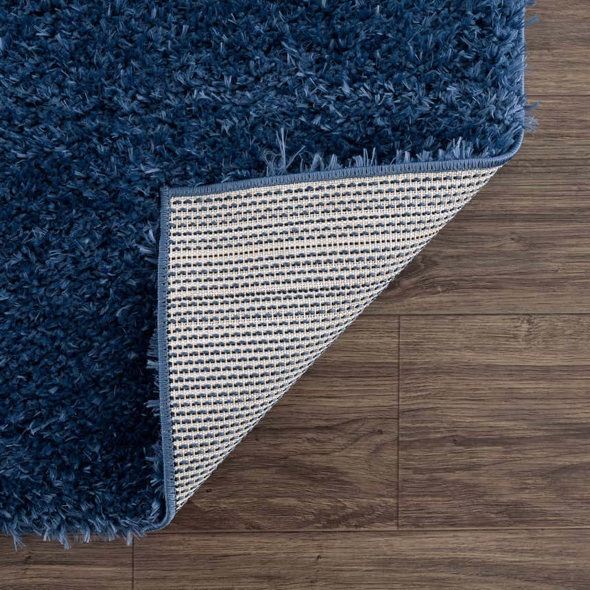 Close-up of the Faina Solid Dark Blue Shag Rug showing its plush texture and backing on a wood floor. Fold