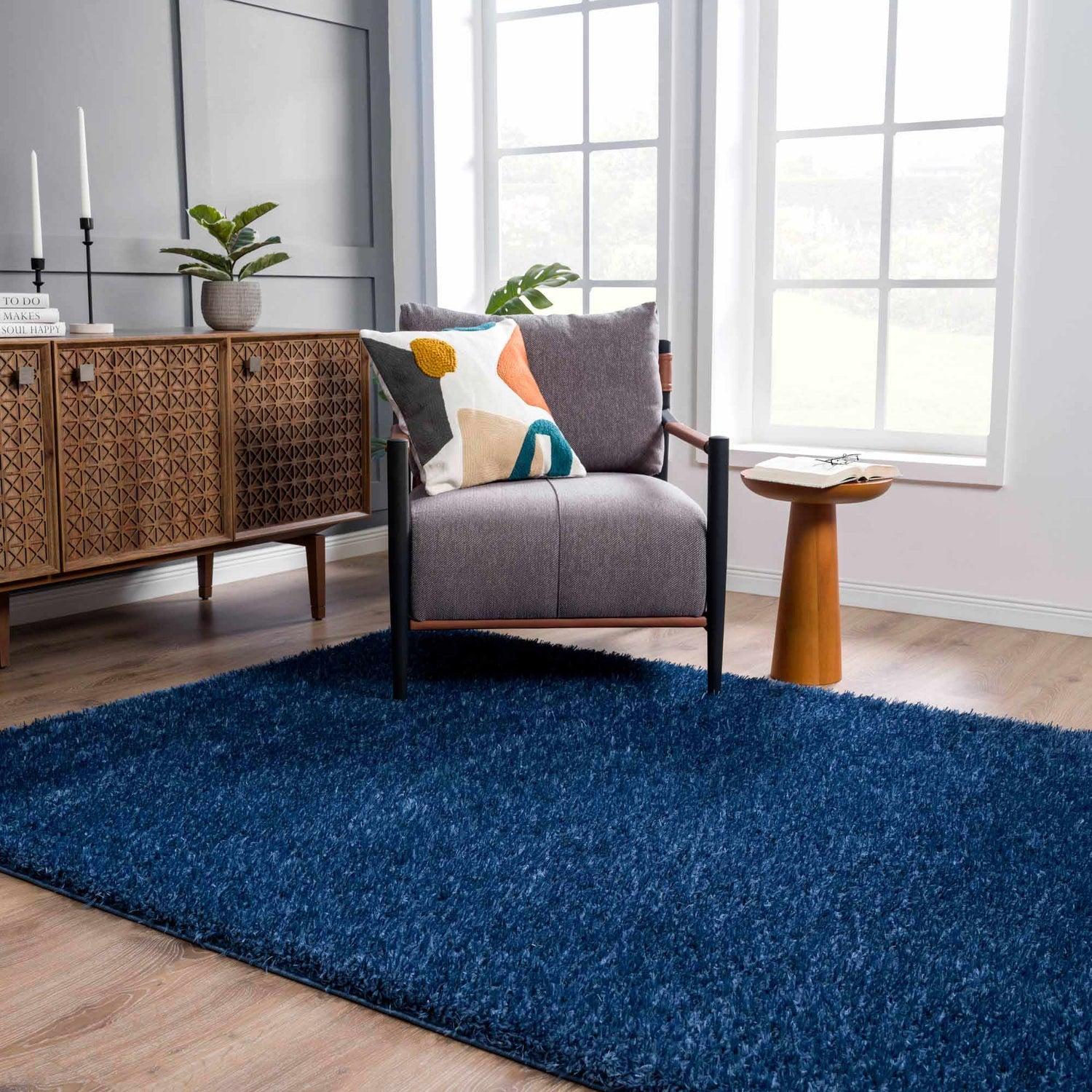 Living room scene with Faina Solid Dark Blue Shag rug, gray chair, patterned pillow, and wooden accent table. Roomscene-Rectangle