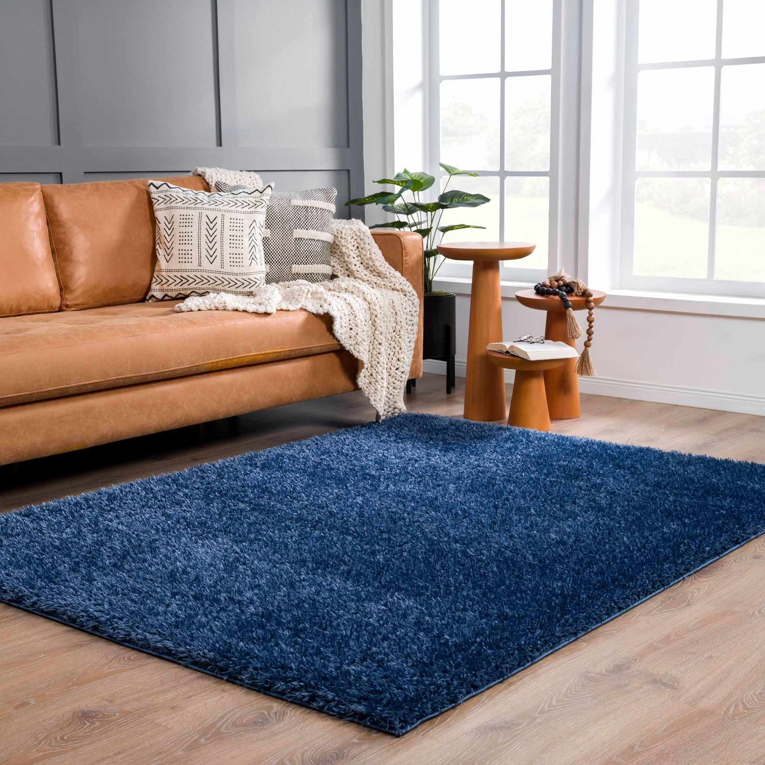 Living room scene with a Faina Solid Dark Blue Shag rug, tan leather couch, wood side tables, and bright natural light. Roomscene-Rectangle