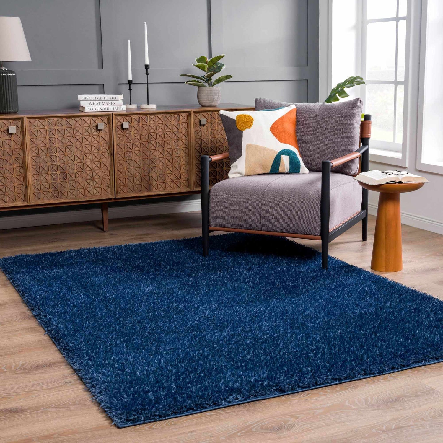 Living room scene with Faina Solid Dark Blue Shag Rug, wood cabinet, gray chair, and light wood floors. Roomscene-Rectangle